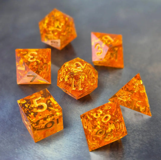 A set of sharp-edge orange resin dice with sparkling gold foil and gold numbers, displayed on a metallic surface.