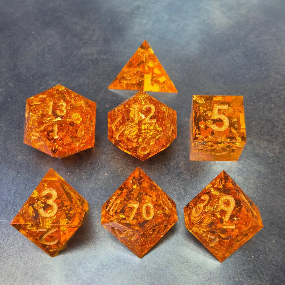 A set of sharp-edge orange resin dice with sparkling gold foil and gold numbers, displayed on a metallic surface.