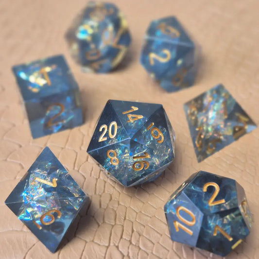 A set of sharp-edge resin dice with ocean-blue transparency, shimmering foil inclusions, and gold numbering, displayed on a textured beige surface.