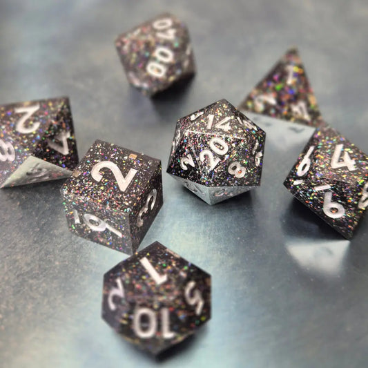 A set of black sharp edge resin dice with white numbers and colourfull sparkles on a blue metal surface