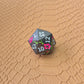A set of black metal dice with white numbers and pink flowers with green stems, displayed on a brown textured surface.